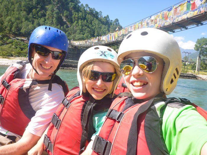 Tim, Tyler and Kara on a raft, smiling at the camera as they float under a thin pedestrian bridge suspended over the water.