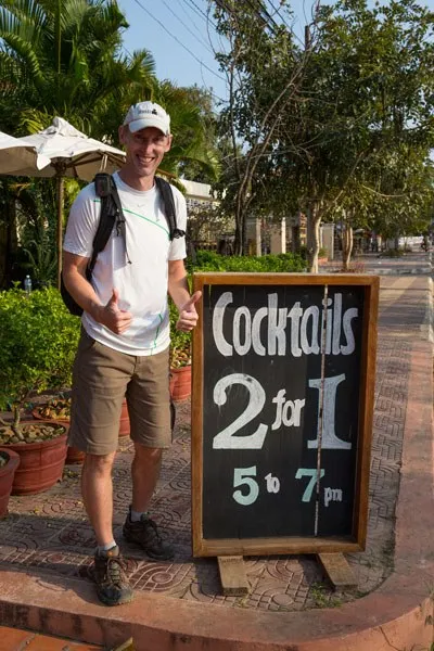 Tim gives a double thumbs up next to a sign reading "Cocktails 2 for 1, 5 to 7pm" at the Rikitikitavi Hotel, Kampot, Cambodia