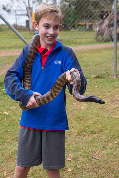 Tyler stands and smiles with a large snake draped around his neck. He holds the snake's upper body with both hands.