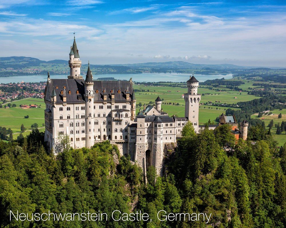 a castle on a hill with trees and a river in the background with Neuschwanstein Castle in the background
